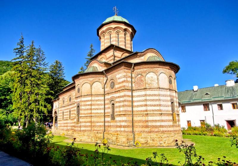 Imagine cu Mănăstirea Cozia, așezată pe malul Oltului, cu arhitectura bizantină specifică secolului XIV și peisaj montan în fundal