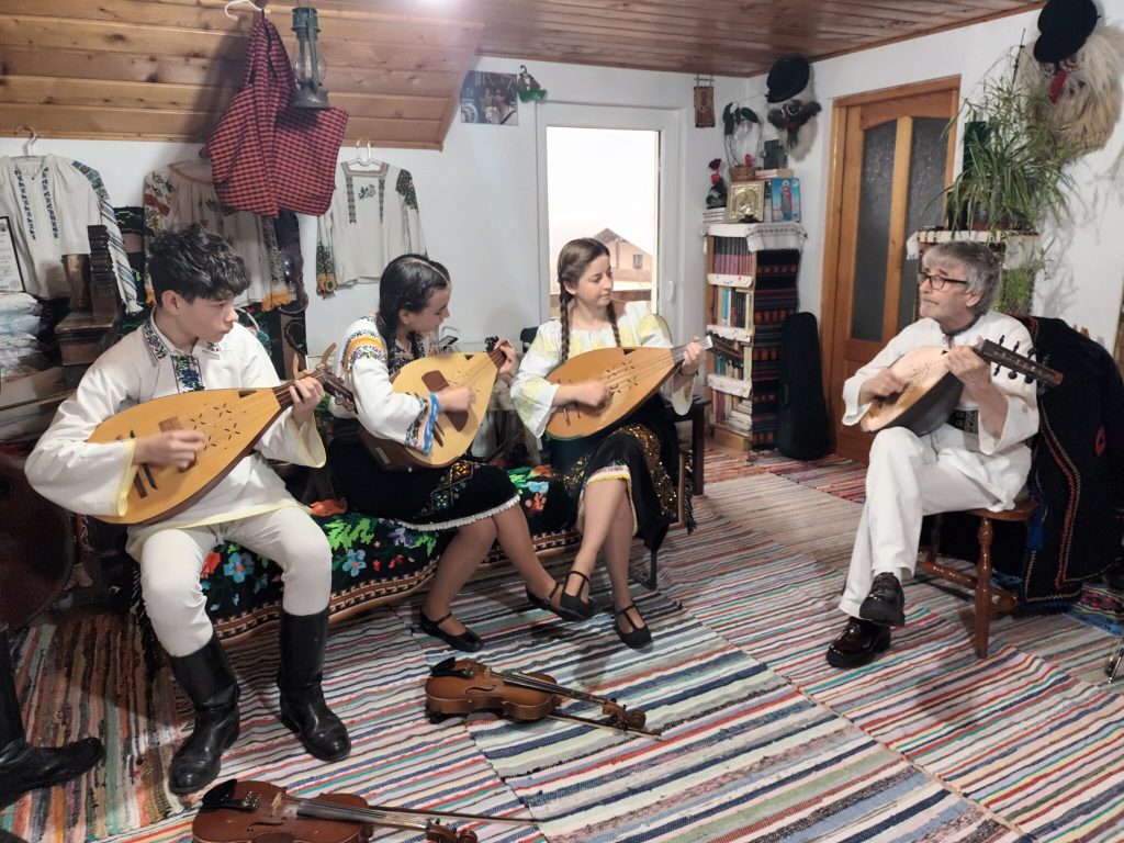 Această imagine captează spiritul viu al tradiției muzicale din zona Balcanilor. Grup de patru muzicieni în costume tradiționale românești cântând la cobză (instrumente cu coarde) într-o cameră decorată cu obiecte de artizanat și ii tradiționale.