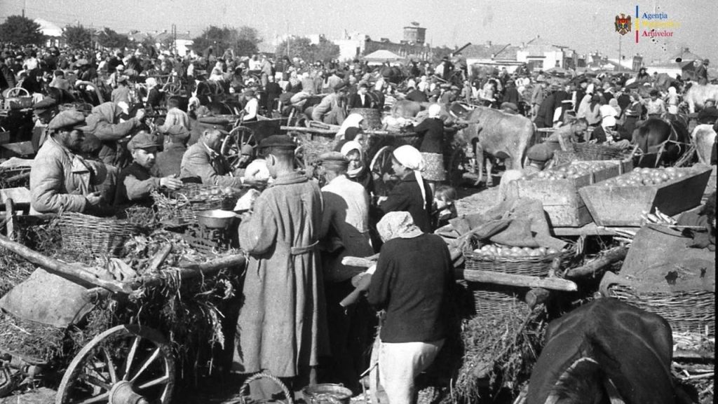 Piață aglomerată în timpul foametei din Moldova, cu țărani, căruțe și produse agricole într-o fotografie istorică alb-negru.